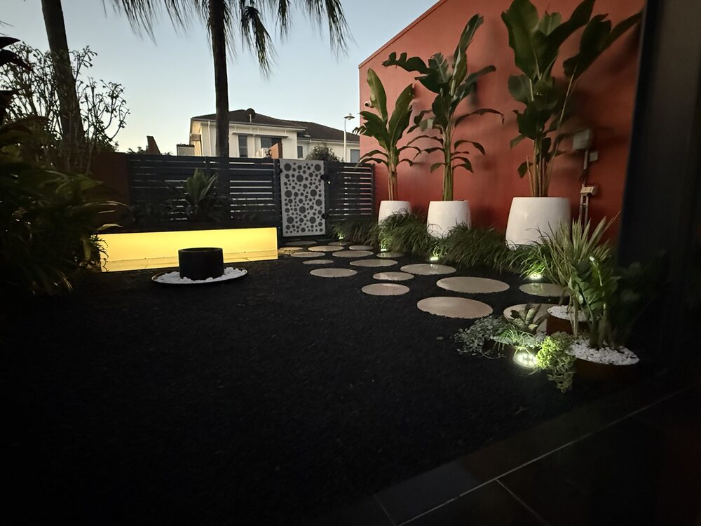 Landscaped courtyard at dusk with stepping stones, an aluminium screen, an uplit planter and feature lighting